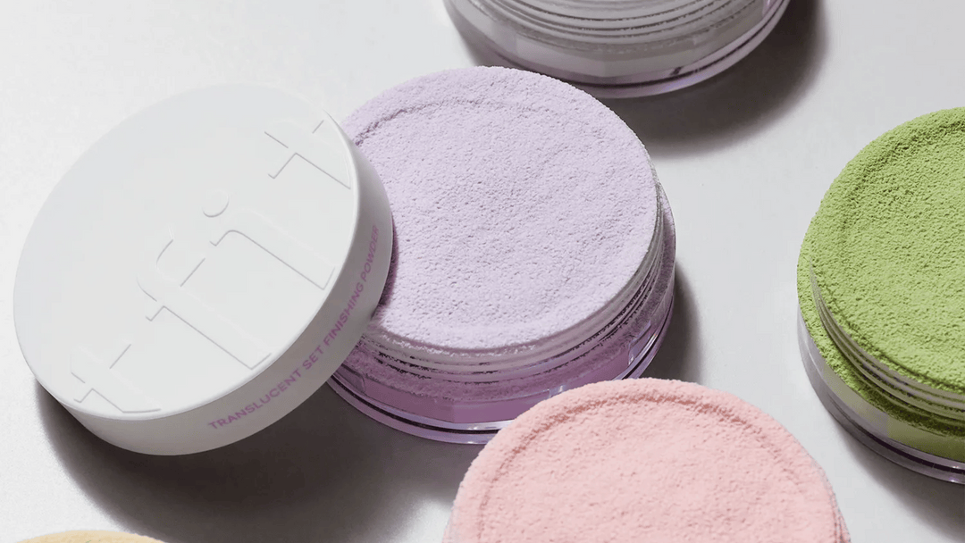 Närbild på olika nyanser av lösa, genomskinliga setting-puder i behållare. Pudren visas i lavendel, rosa och gröna nyanser, där lavendelpudret är i förgrunden.