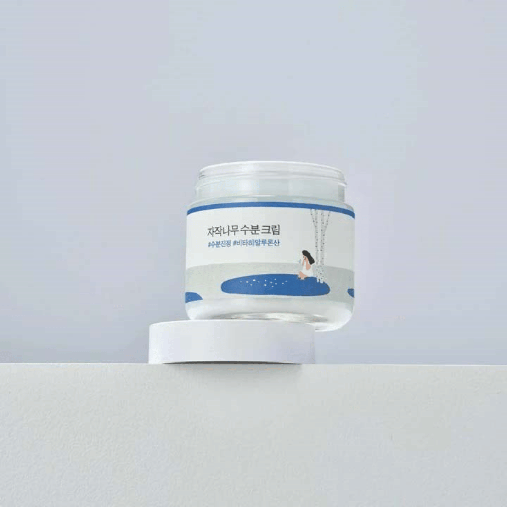 Round Lab Birch Juice Moisturizing Cream i transparent burk med vit etikett och blå illustration av kvinna vid björk, stående på vit yta mot ljus bakgrund.