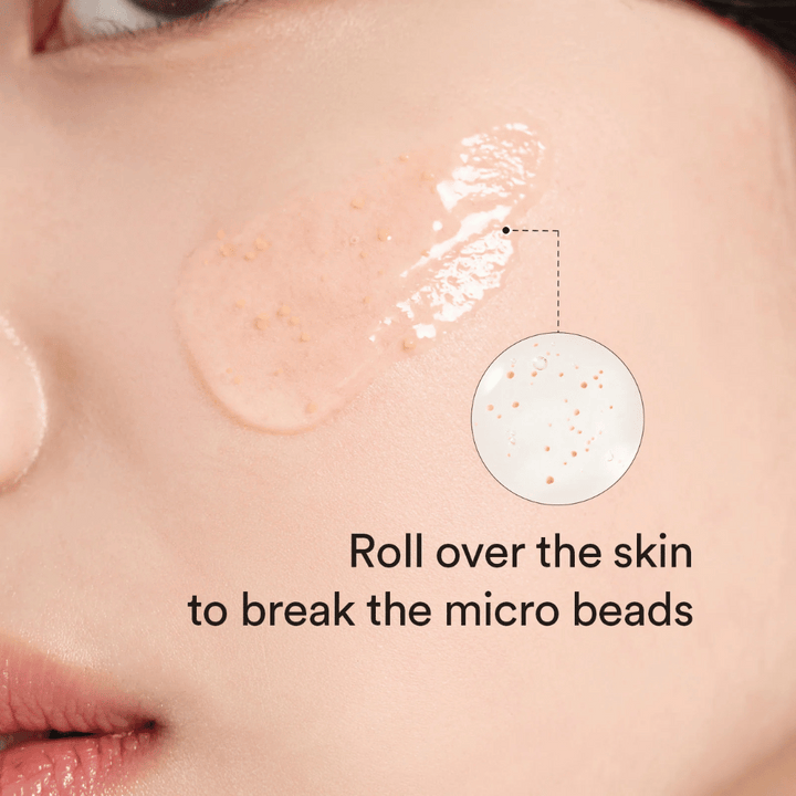 Närbild på en kind med ett serum innehållande små mikropärlor applicerat på huden. En cirkelzoom visar pärlorna tydligare. Texten säger “Roll over the skin to break the micro beads”.