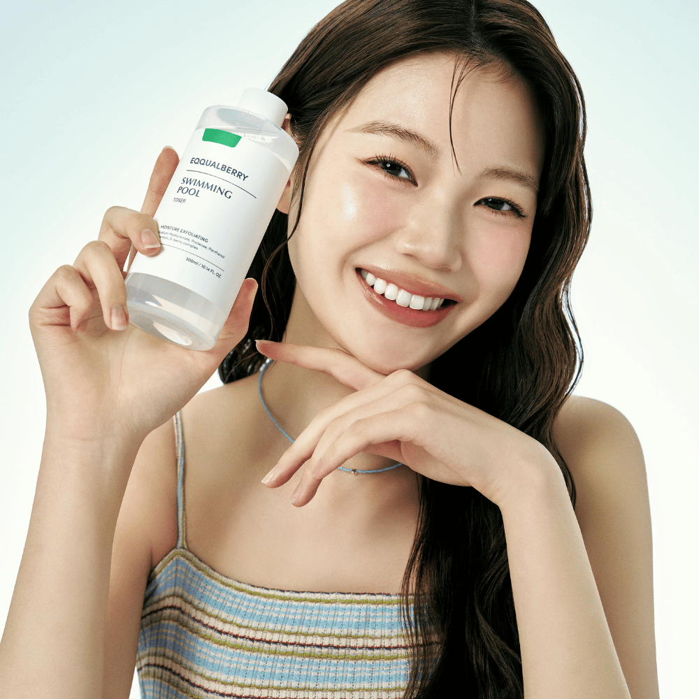 Leende kvinna med strålande hud håller upp Equalberry Swimming Pool Toner. Fräsch och naturlig look med lätt smink och en somrig outfit i blå toner.
