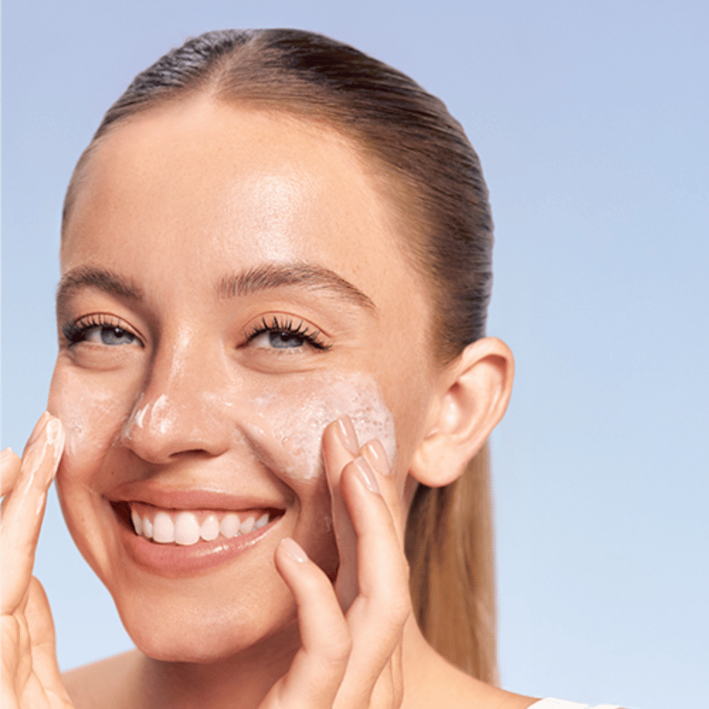 Leende kvinna applicerar LANEIGE Water Bank Gentle Gel Cleanser i löddrande form på kinderna, mot blå bakgrund.