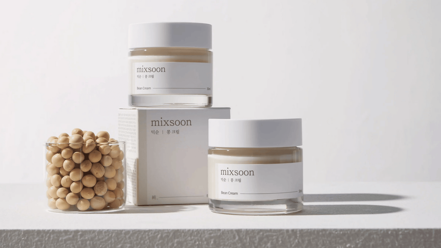 Två burkar av Mixsoon Bean Cream står på en ljus yta med en glasbehållare fylld med beige bönor och en produktkartong bredvid. Enkel och ren design med tydliga etiketter. Bakgrunden är neutral för att fokusera på produkterna.