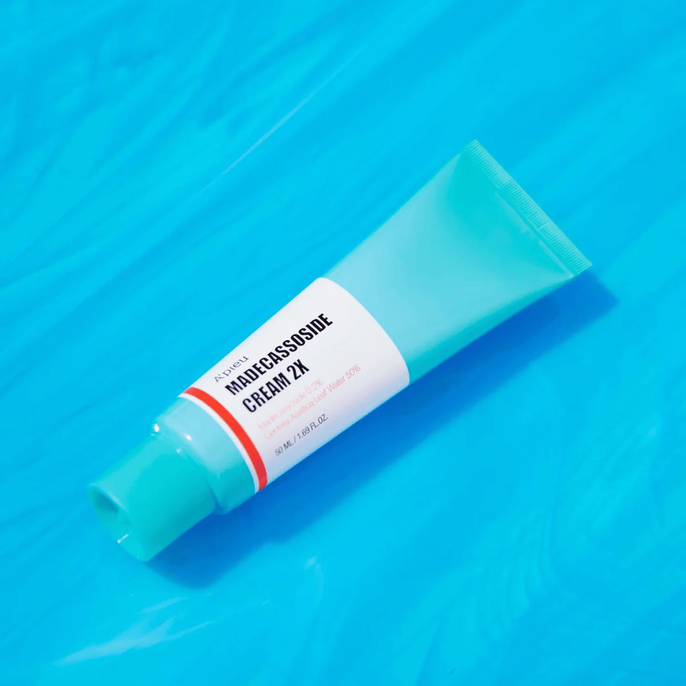 En tub A'PIEU Madecassoside Cream 2X ligger diagonalt över en blå bakgrund med vattenliknande vågor.