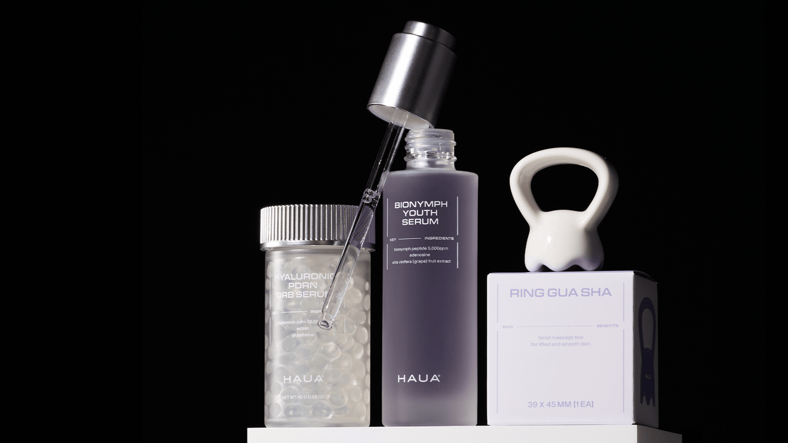 HAUA Bionymph Youth Serum, Hyaluronic PDRN Orb Serum och Ring Gua Sha uppställda mot svart bakgrund. Serumflaskan med pipett är öppen och omges av hudvårdsprodukter och massageverktyg i en stilren premiumkomposition.