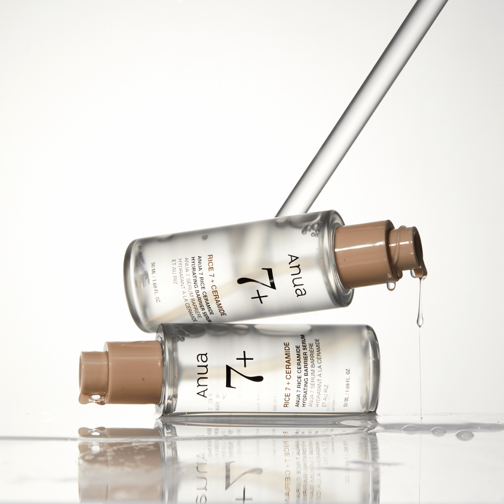 The image shows two transparent bottles of "Anua 77 Rice + Ceramide Hydrating Barrier Serum" with drops running from a pipette. The serum, enriched with rice and ceramides, is intended to hydrate and strengthen the skin's barrier. The bottles are elegantly designed, enhancing the product's premium feel.
