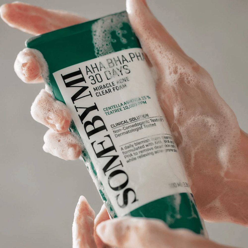 A person holds a tube of "SOME BY MI AHA, BHA, PHA 30 Days Miracle Acne Clear Foam" with foaming content over the hands. The product is intended for daily use to combat acne and cleanse pores with its active ingredients such as centella, tea tree oil, and acids like AHA, BHA, and PHA.