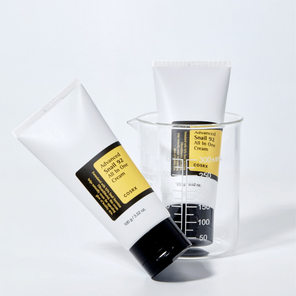 A tube of COSRX Advanced Snail 92 All in One Cream lying next to an upside-down transparent measuring cylinder on a white background.