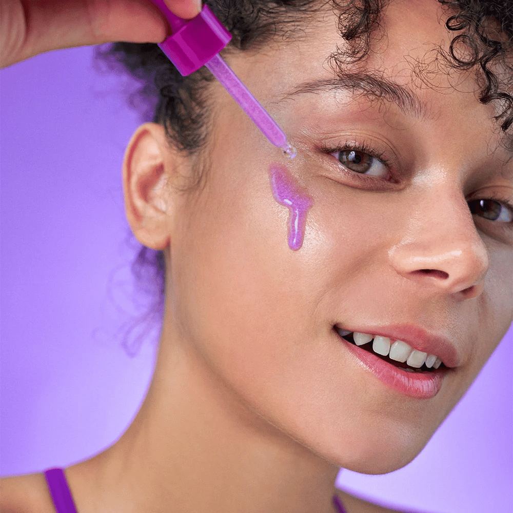 Smiling woman applies a purple shimmering serum to the skin with a pipette. Radiant complexion and purple background matching the product's color tone.