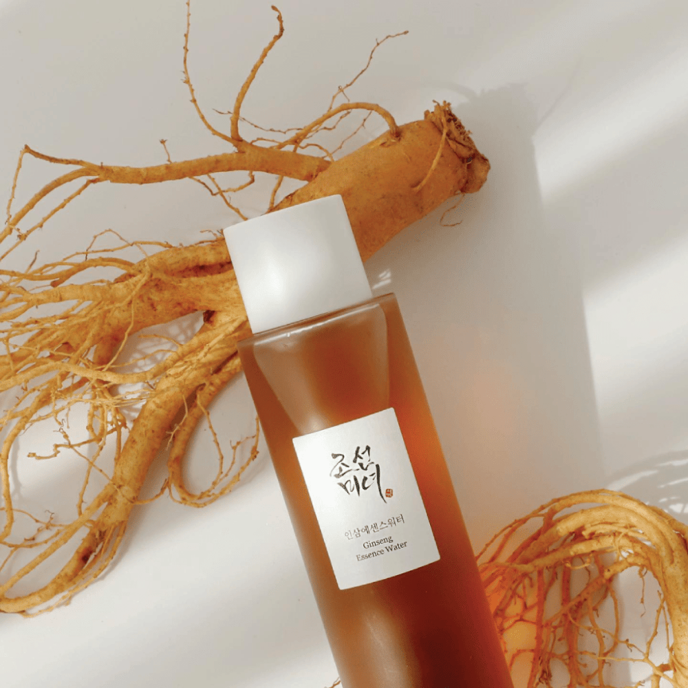 En brun flaska Beauty of Joseon Ginseng Essence Water med vit kork ligger bredvid en färsk ginsengrot på vit bakgrund.