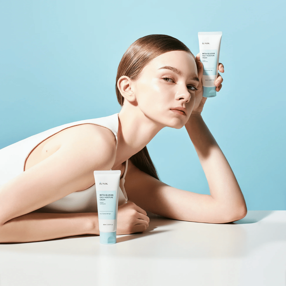 Lifestyle image in which a model poses with iUNIK Beta-Glucan Daily Moisture Cream against a light blue background. The image conveys a clean, fresh, and moisturizing feel while clearly showcasing the product.
