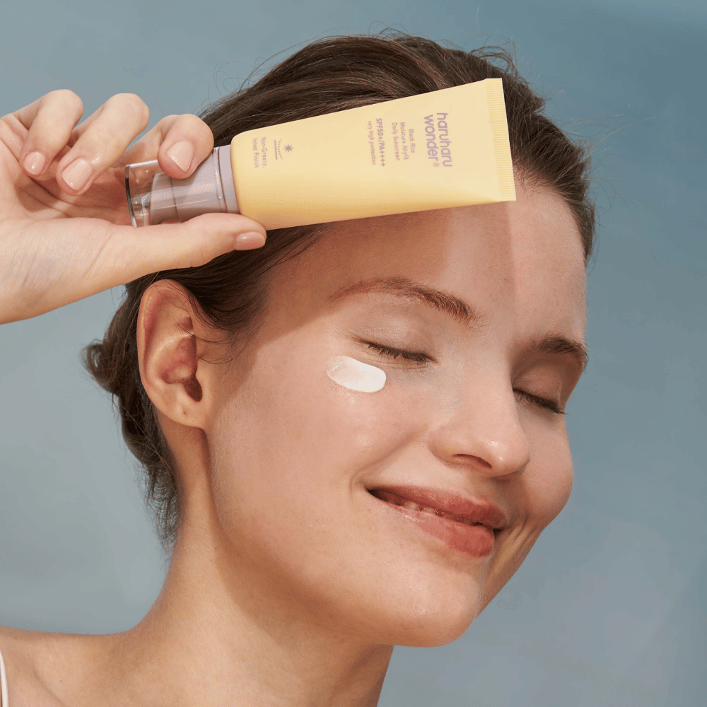 A smiling woman applies Black Rice Moisture Airyfit Daily Sunscreen SPF 50+ PA++++ from Haruharu Wonder on her face. She holds a yellow tube of sunscreen and has a dab of the product on her cheek. The background is light blue.