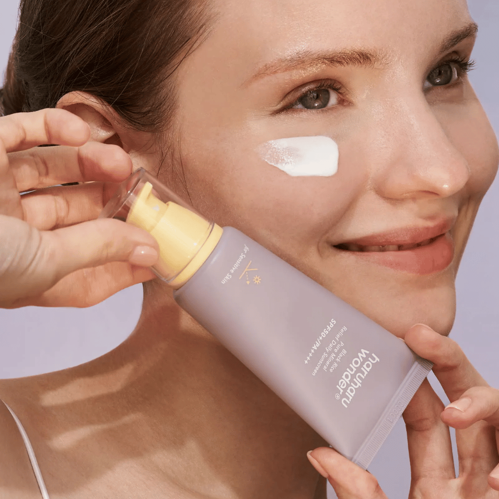 A smiling woman applies Haruharu Wonder Black Rice Pure Mineral Relief Daily Sunscreen SPF 50+ PA++++ on her face. She holds a purple tube of sunscreen and has a dab of the product on her cheek. The background is light purple.
