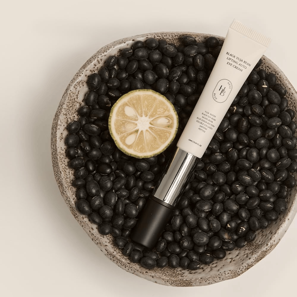 Black Yuja Bean Lifting Auto Eye Cream lying in a bowl filled with black beans and a halved citrus fruit – a visual representation of the product's natural ingredients.