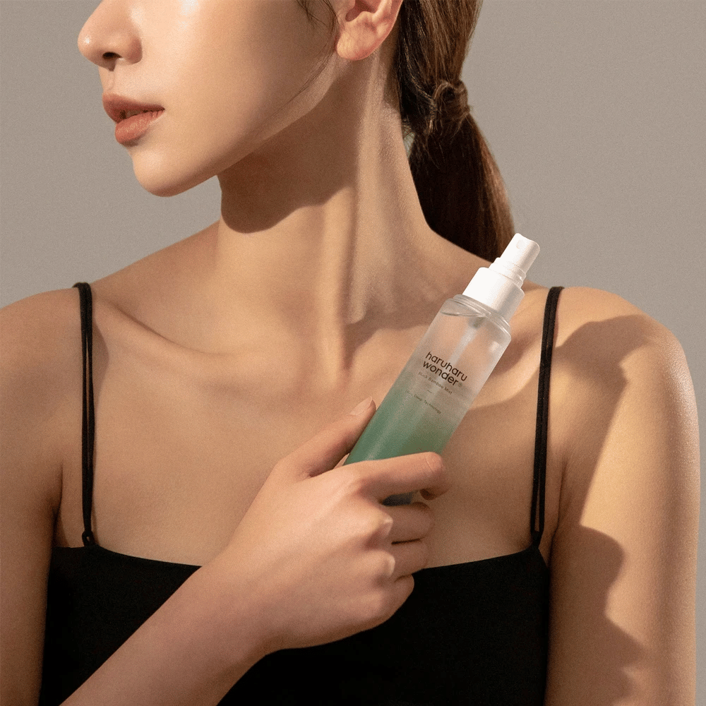 Image of HaruHaru Wonder Black Bamboo Mist held by a woman. The product is in a transparent spray bottle with a green tint and a white pump. The woman has a relaxed look, wears a black top, and focuses on using the mist to give her skin moisture and radiance. The bottle is part of a skincare routine.