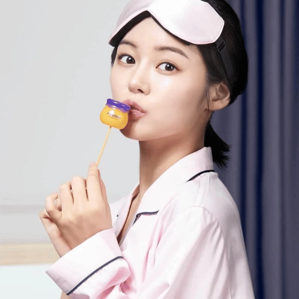 The woman in the picture is using a blueberry honey lip balm from FRUDIA, suggesting a skincare routine with natural ingredients.