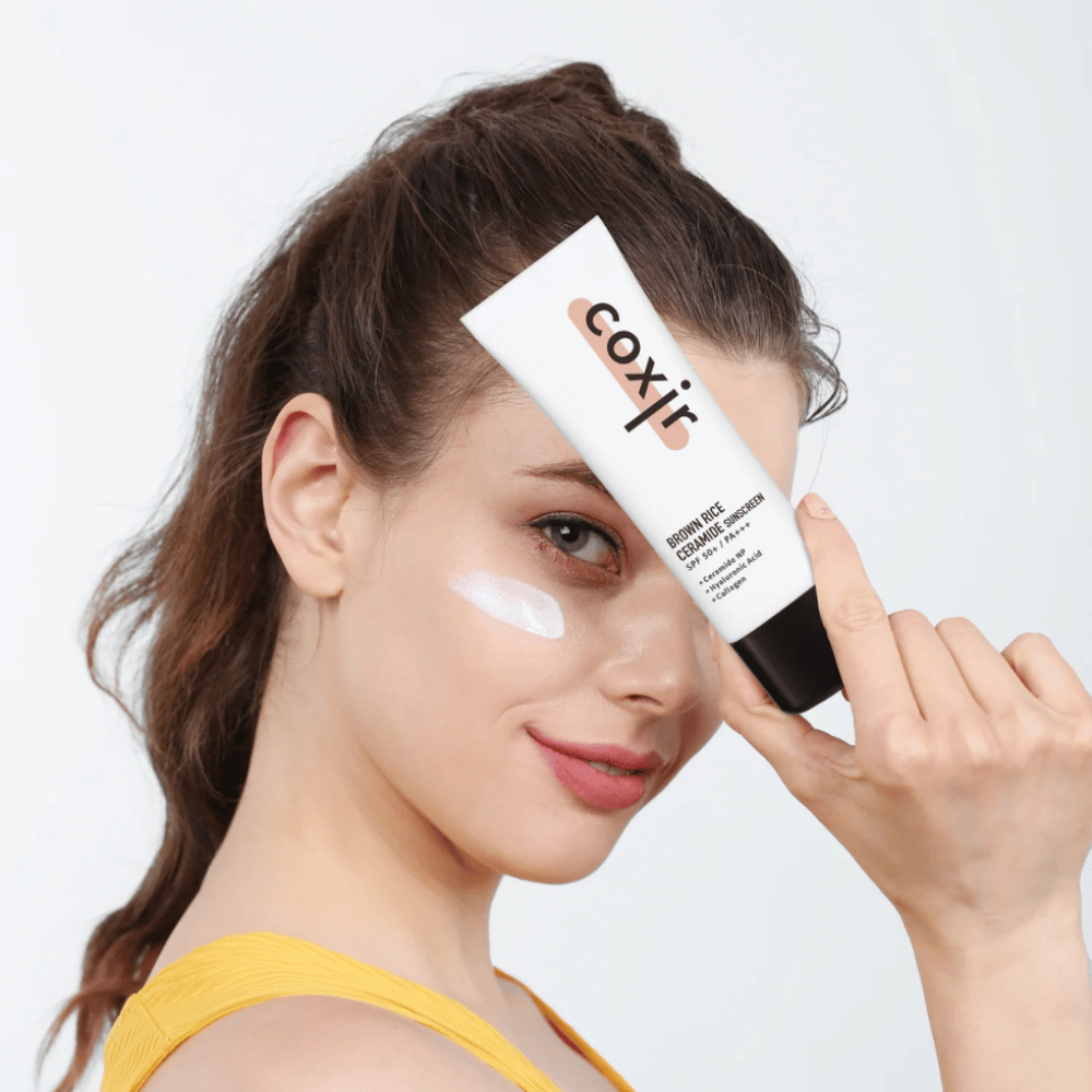 Woman with sunscreen applied on her cheek holds up a tube Coxir Brown Rice Ceramide Sunscreen, SPF 50+ PA+++, towards the forehead and smiles slightly.