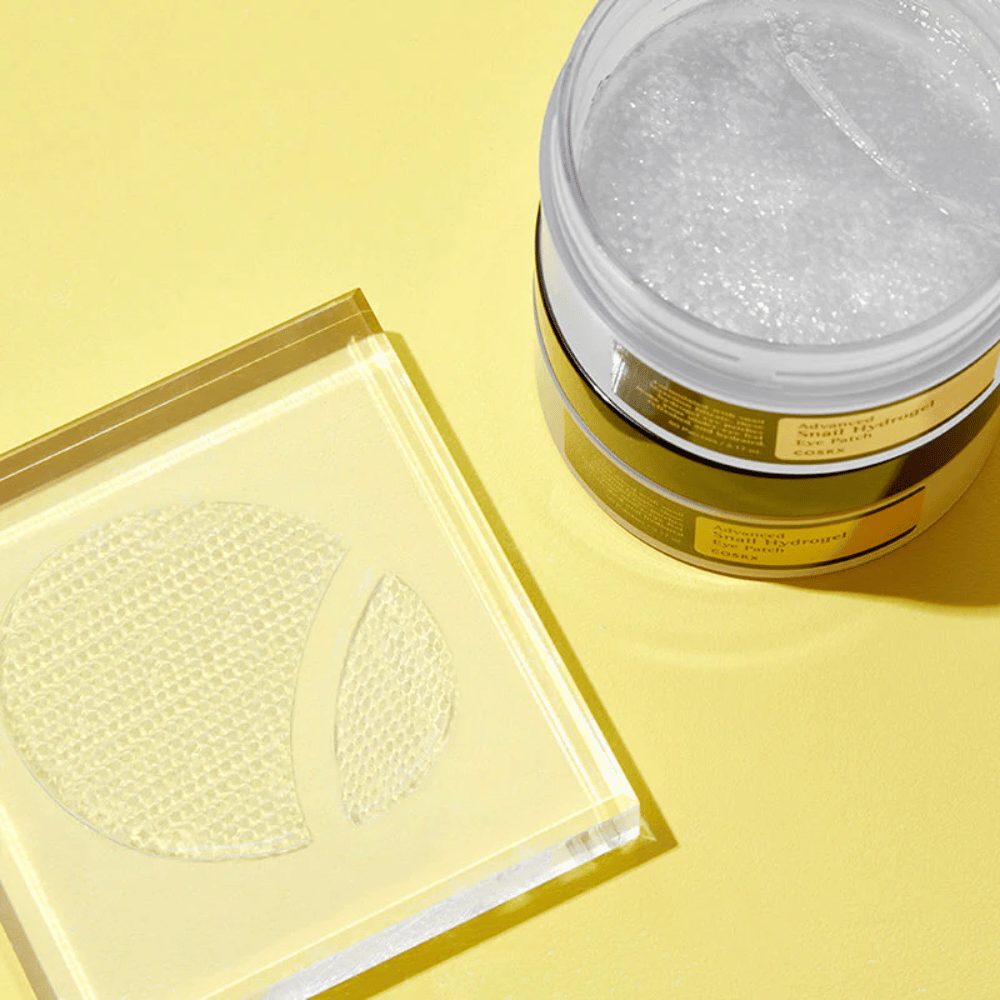 An open jar COSRX Advanced Snail Hydrogel Eye Patch stands on a yellow background next to a transparent coaster where two eye patches are placed.