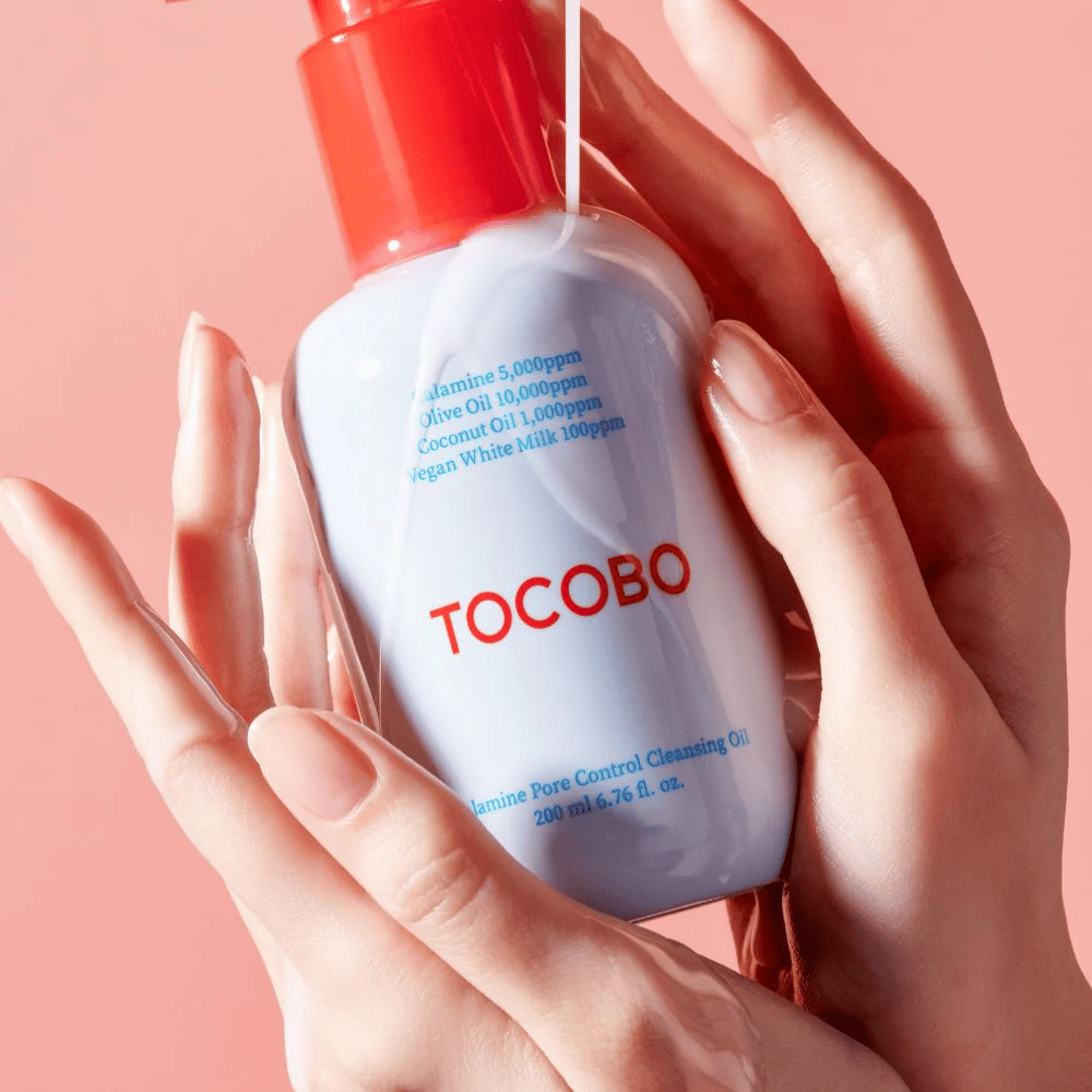 TOCOBO Calamine Pore Control Cleansing Oil held in hands against a pink background, the bottle's red pump and ingredient list are clearly visible.