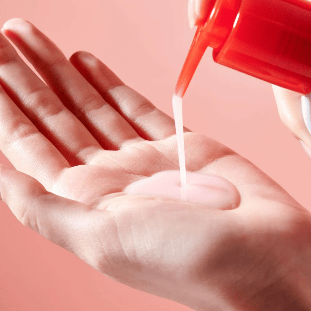 A light pink cleansing oil is dispensed from a red pump bottle into an open hand against a pink background.