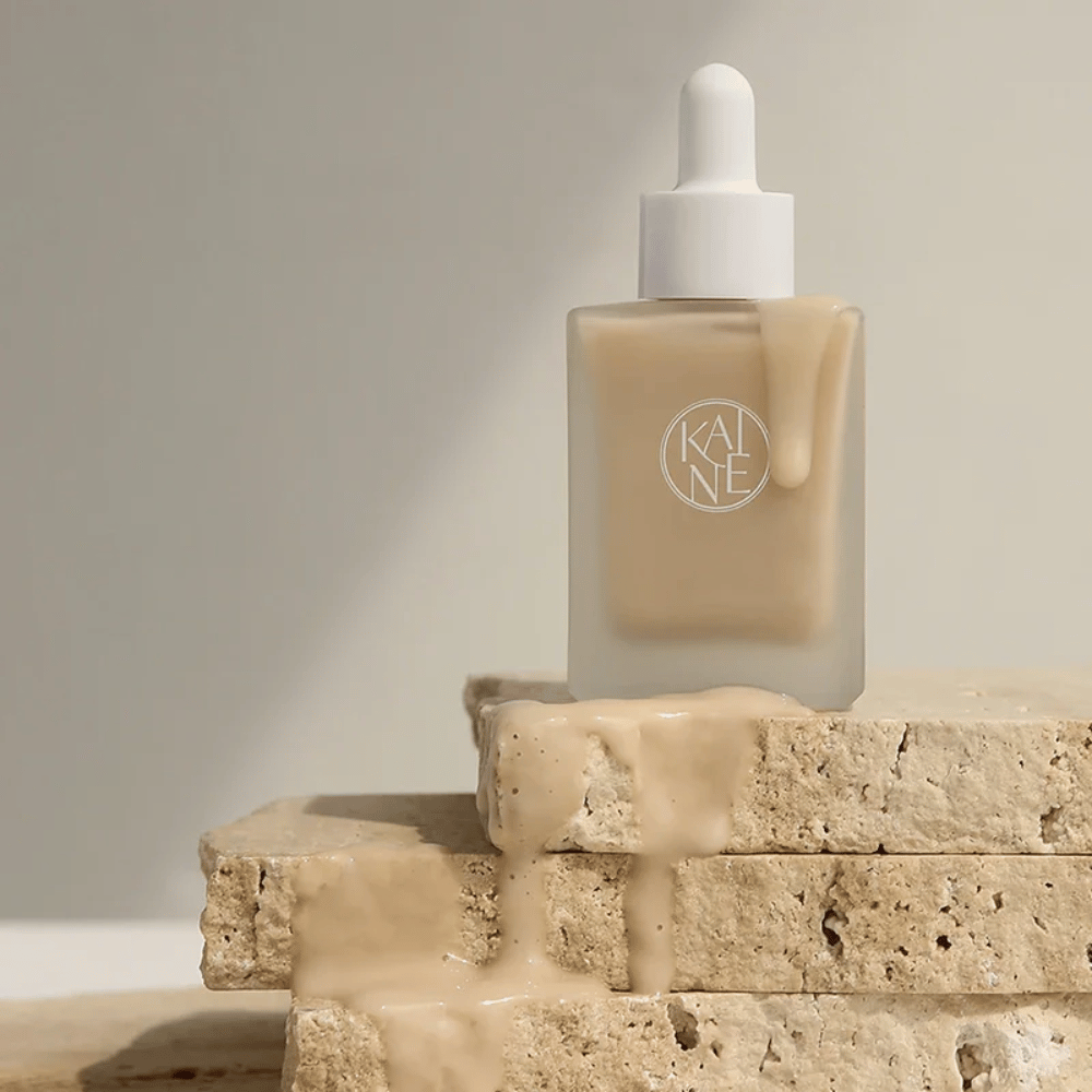 KANE Chaga Collagen Charging Serum in a frosted glass bottle with a dropper. Creamy texture flows over natural stones in a soft and minimalist light.