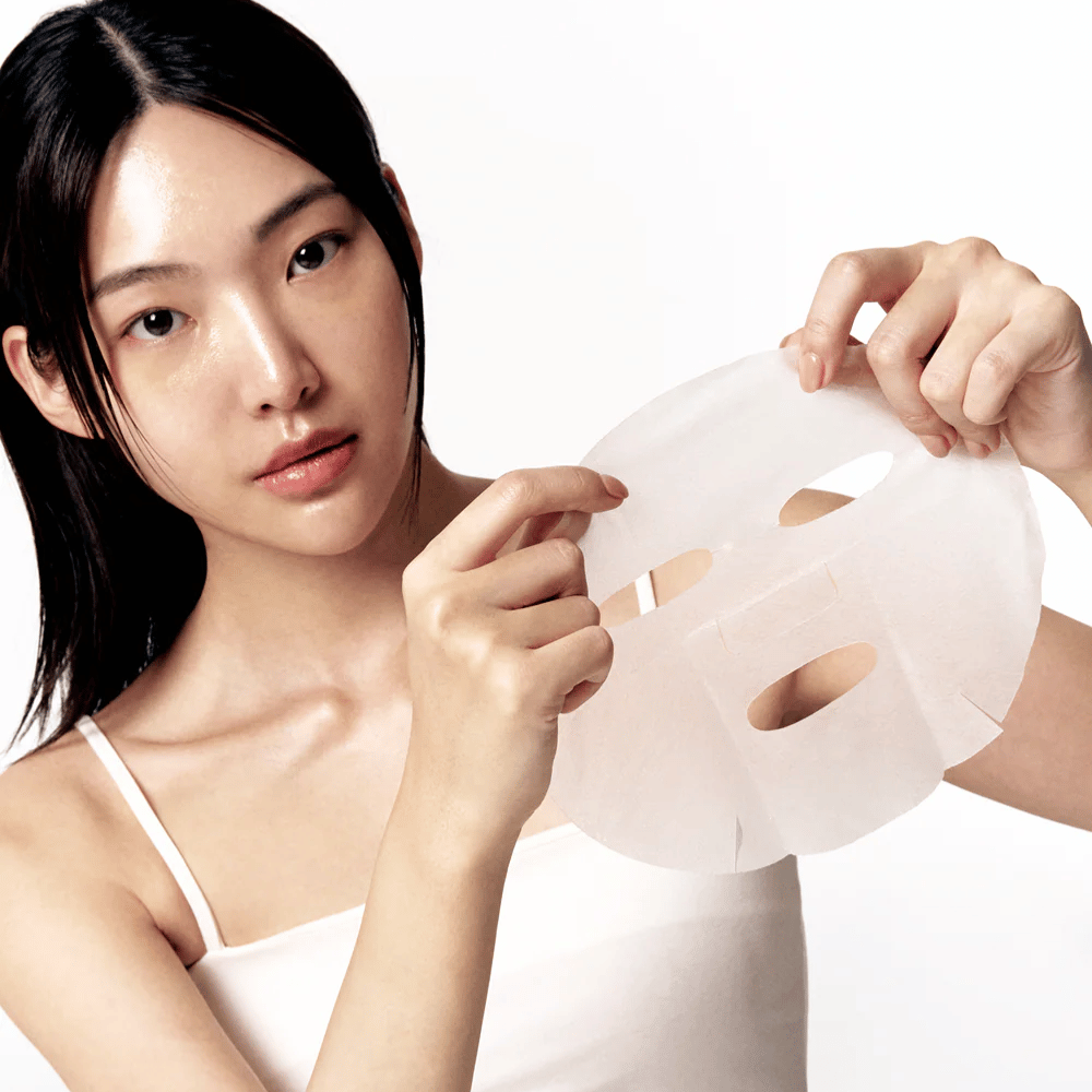 Woman with clean and moisturized skin holds up a white Sheet mask in front of her, ready to apply it to her face.