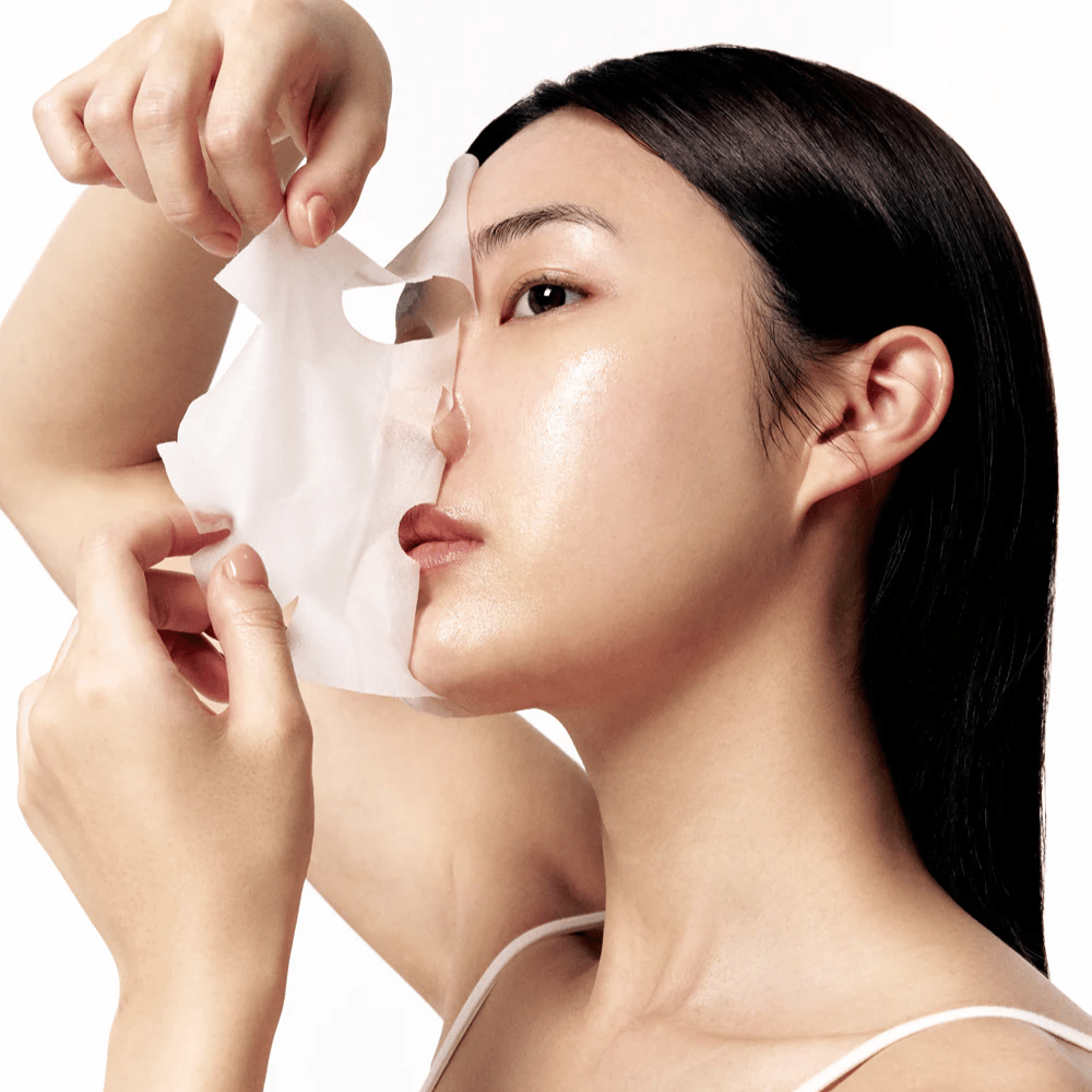 Woman with smooth, moisturized skin removes a white Sheet mask from her face, revealing a radiant and even skin tone.