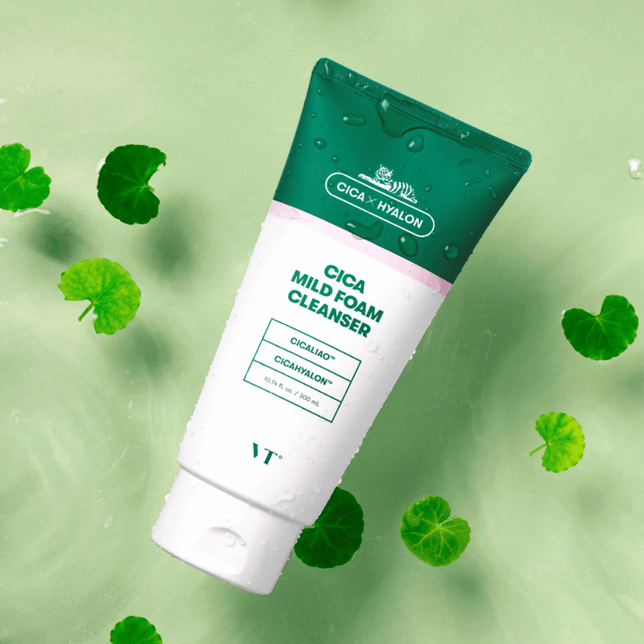 VT Cica Mild Foam Cleanser liggande mot ett ljusgrönt underlag med vattendroppar på förpackningen och omgiven av gröna centella asiatica-blad, vilket framhäver produktens lugnande egenskaper.