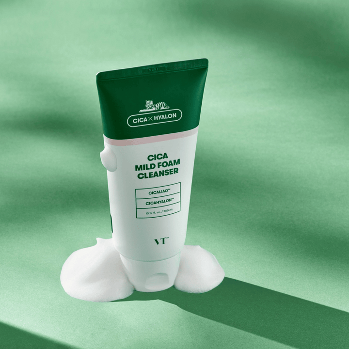 Tub med VT Cica Mild Foam Cleanser stående på ett grönt underlag med lödder runt basen. Produkten har grön och vit design och marknadsförs med CICALIAO™ och CICAHYALON™.
