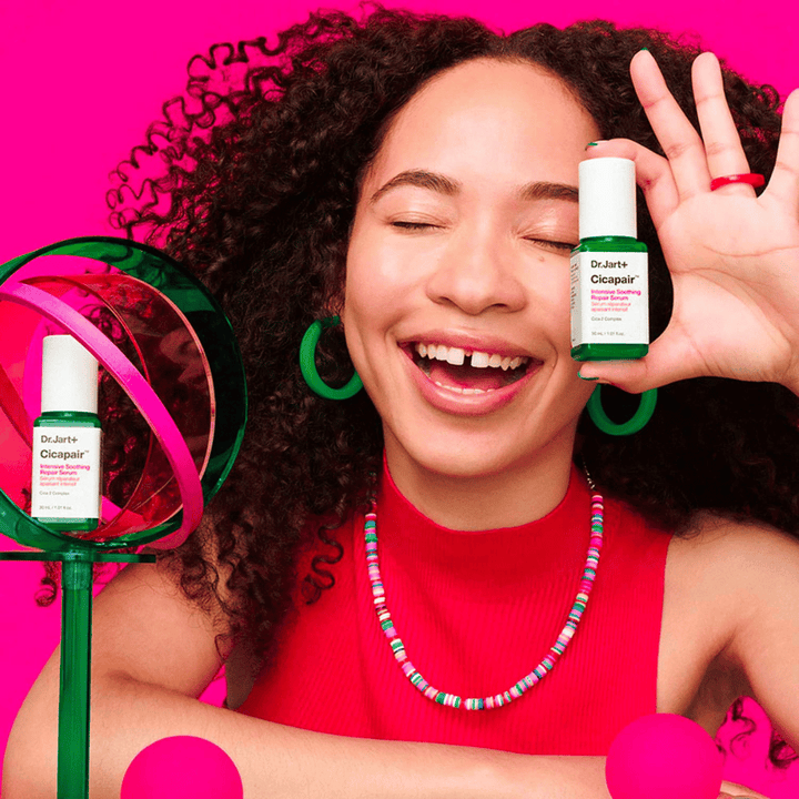 En kvinna ler brett medan hon håller Dr.Jart+ Cicapair Intensive Soothing Repair Serum framför sitt ansikte. Hon bär gröna örhängen och ett rött halsband. Bakgrunden är ljusrosa och dekorerad med gröna och rosa cirklar.