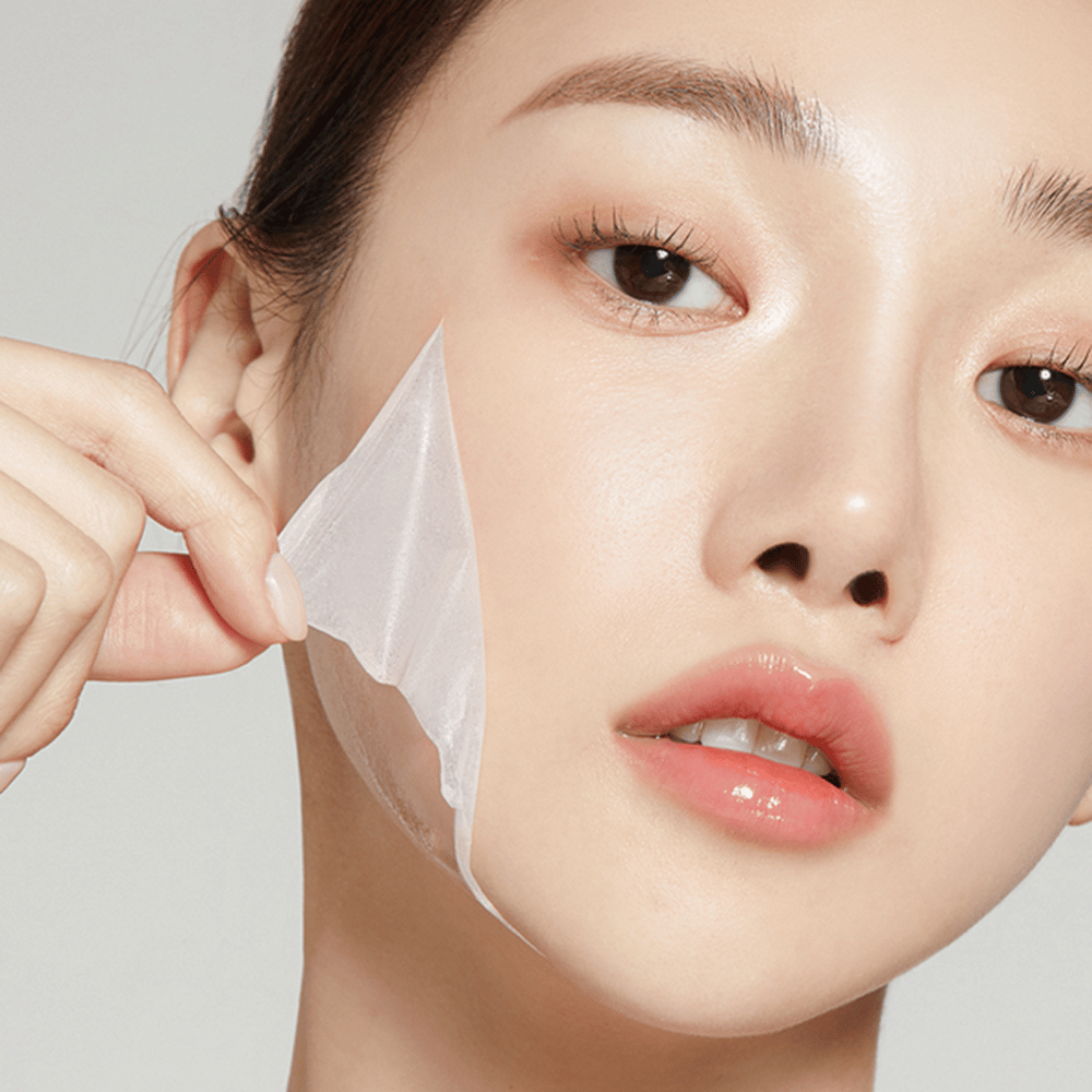 The image shows a young woman removing a thin facial mask, highlighting her hydrated and soft skin. The mask follows the contours of the face, giving the impression of an effective and gentle skincare product. Her expression is relaxed.