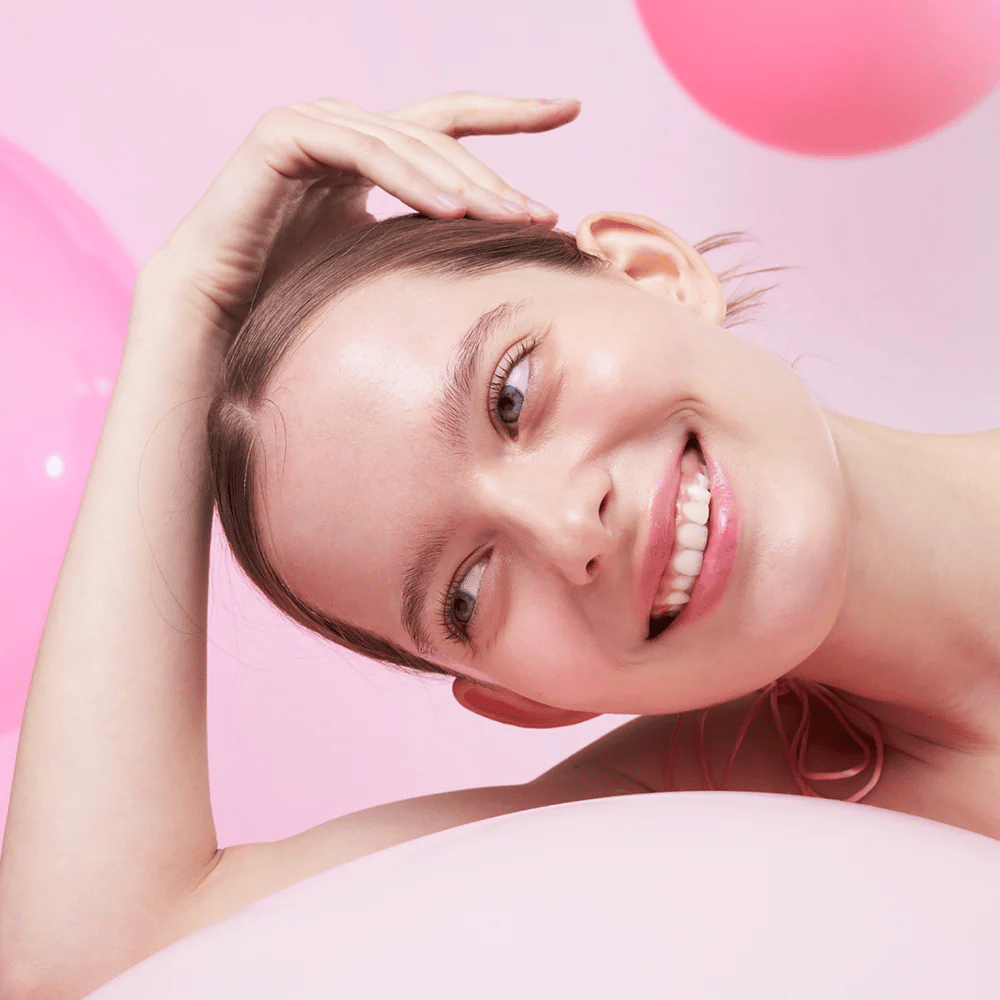 A woman smiles and rests her head on her hand, surrounded by pink balloons. Her skin looks radiant and naturally healthy.