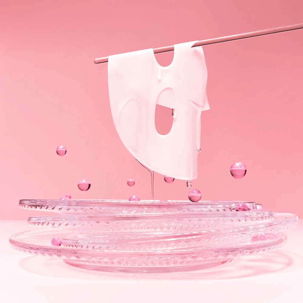 A hydrogel mask hangs on a rod, dripping with serum. The background is pink and the mirror at the bottom reflects the pink bubbles surrounding the mask.