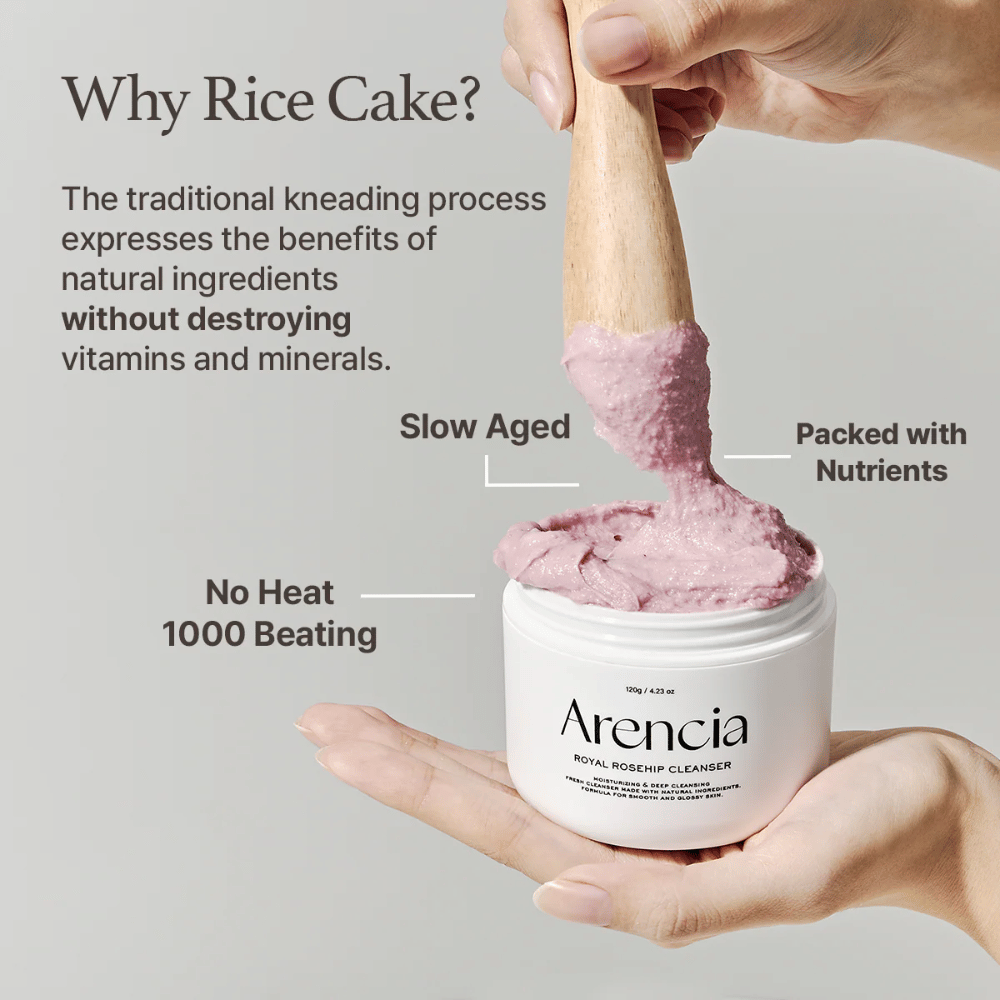 A hand holds an open jar of Arencia Royal Rosehip Cleanser, with a pink, mochi-like cleansing formula being lifted with a wooden spatula – showing the product's texture and traditional manufacturing method.