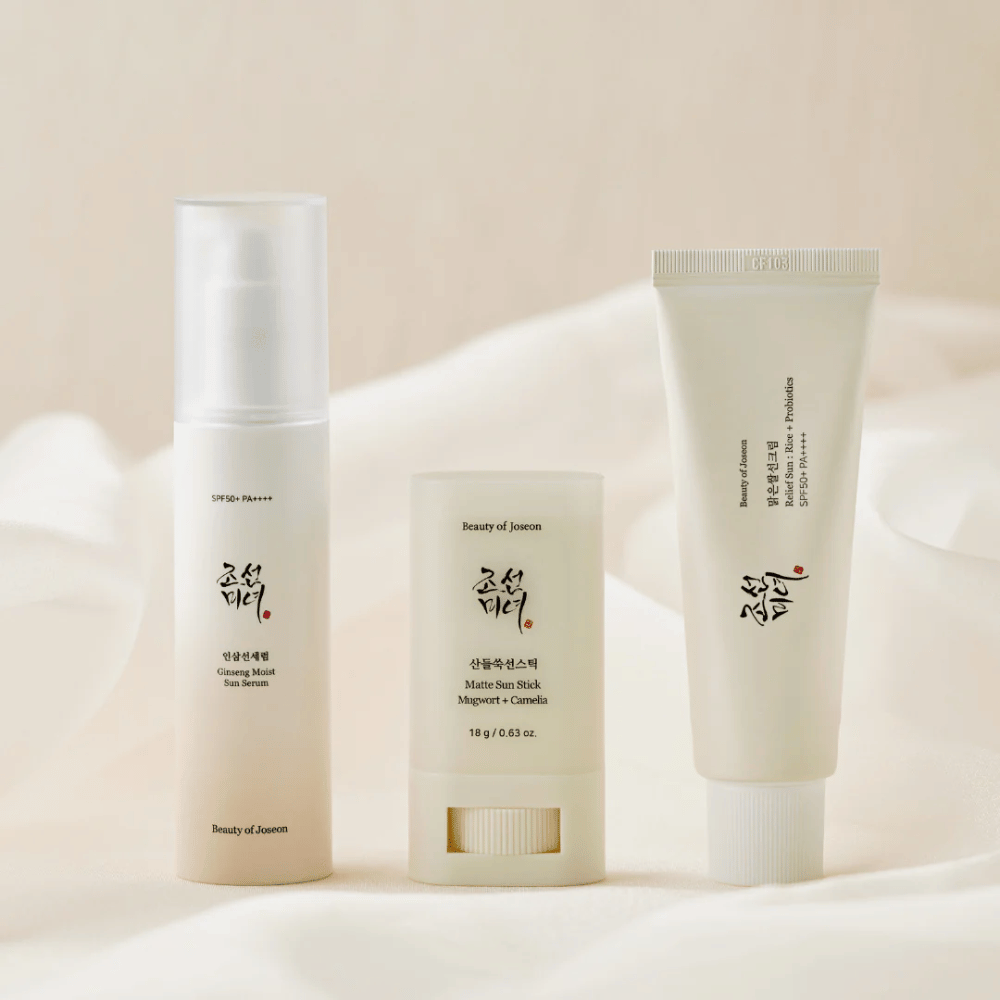 Three sun protection products from Beauty of Joseon - Ginseng Moist Sun Serum, Matte Sun Stick and Relief Sun Rice + Probiotics - are lined up on a cream-colored fabric with soft light.
