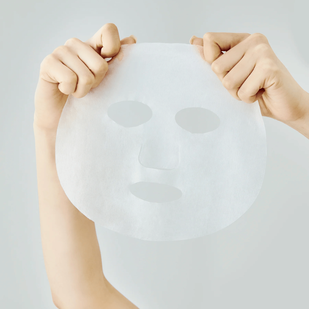 Two hands holding up a white sheet mask against a light gray background, with visible cutouts for eyes, nose, and mouth.