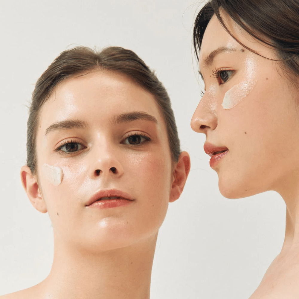 Two women with face masks applied to parts of their faces. One woman has a mask on her cheek and nose, while the other has it under the eye and on the cheek. They have natural makeup and look fresh, highlighting the moisturizing and brightening effect of the masks.