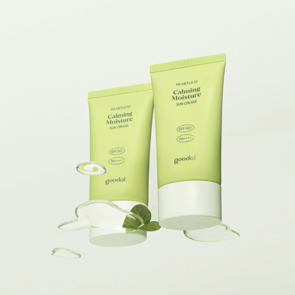 Two tubes of Goodal Heartleaf Calming Moisture Sun Cream SPF50+ PA++++ surrounded by plant leaves and serum drops against a light background.