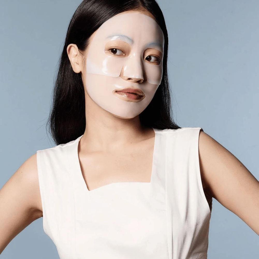 A woman in a white sleeveless dress wears a transparent face mask that covers almost her entire face. The mask gives an impression of advanced skincare and provides the appearance of a beauty treatment, designed to deeply hydrate and treat the skin.