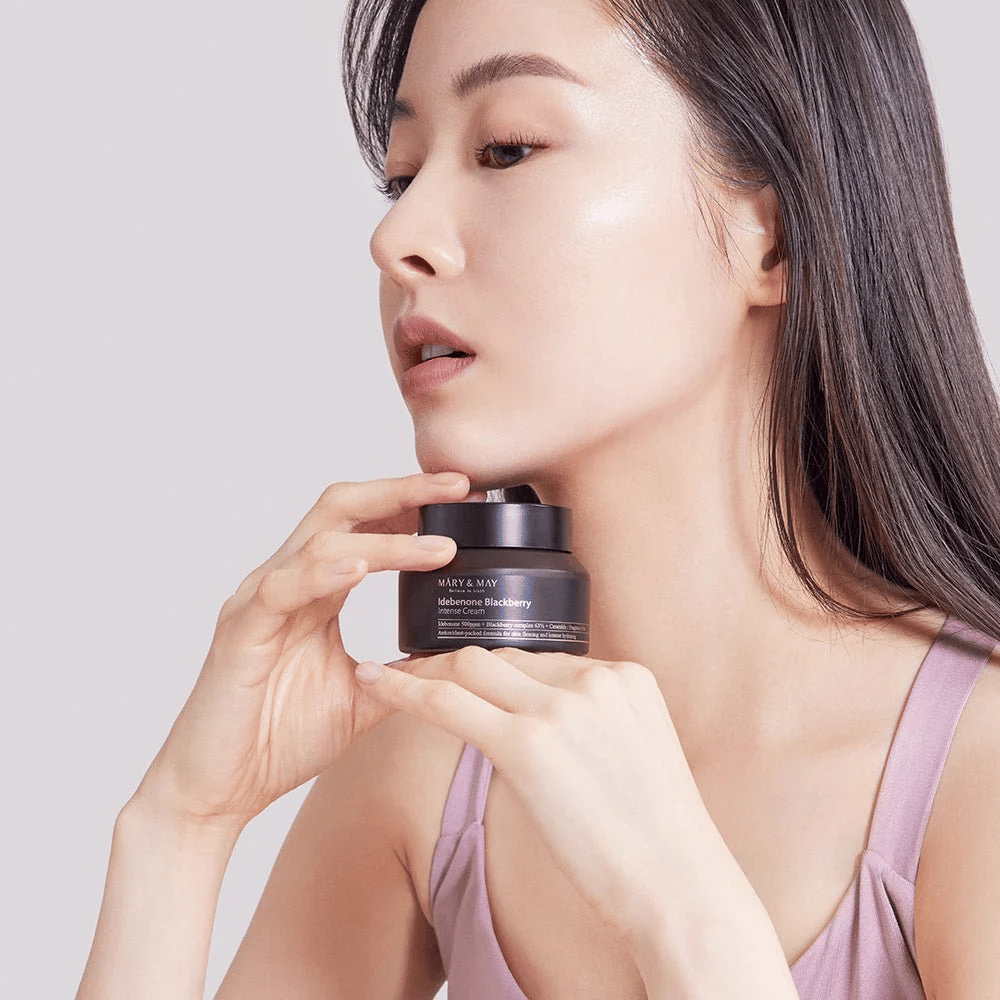 A woman holds a jar of MARY & MAY Idebenone Blackberry Intense Cream. She has radiant skin and smiles softly, dressed in a sleeveless top. The product is an intensive cream with idebenone and blackberry complex intended for skin firmness and hydration.