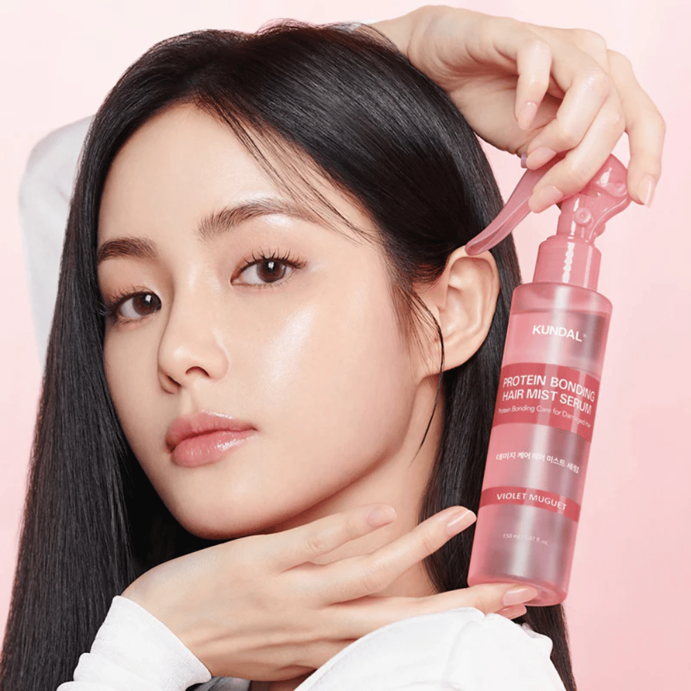 Woman with long black hair holding KUNDAL Protein Bonding Hair Mist Serum Violet Muguet in front of her face, against a light pink background.
