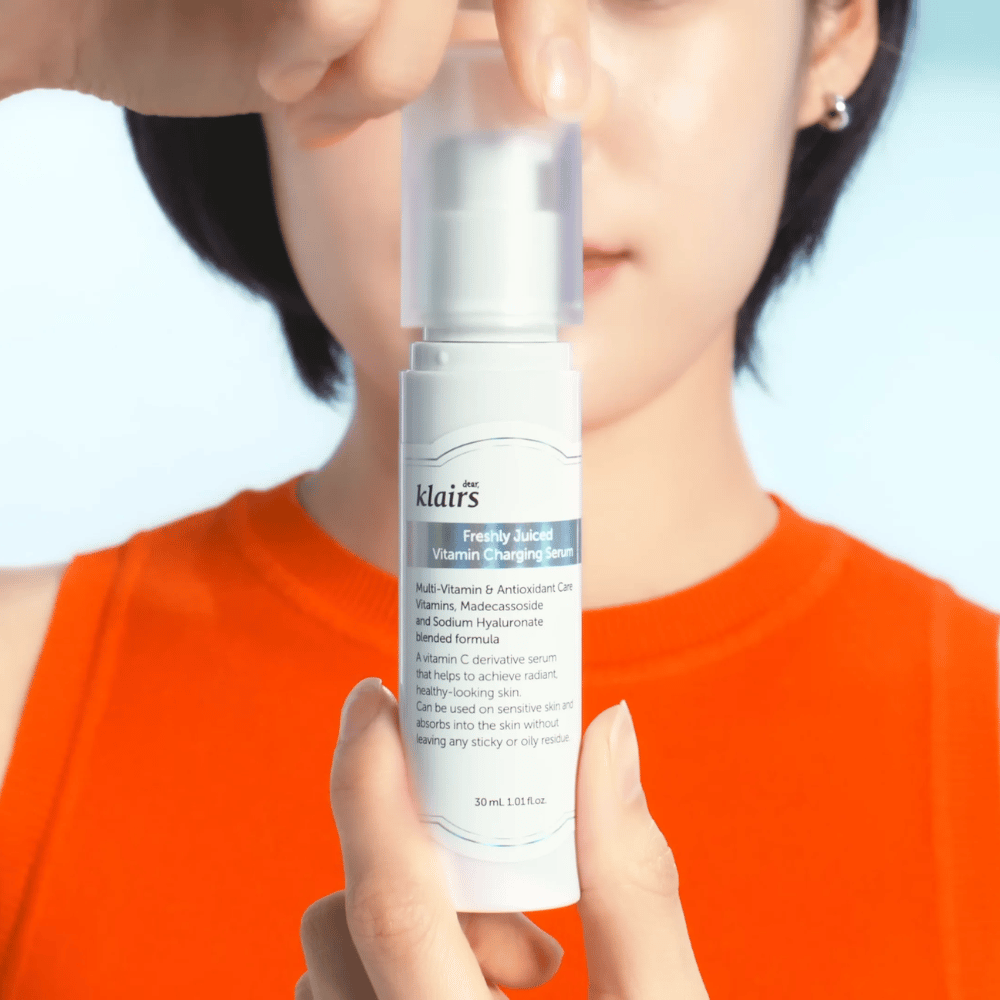Woman in an orange shirt holds Klairs Freshly Juiced Vitamin Charging Serum in front of her against a blue background.