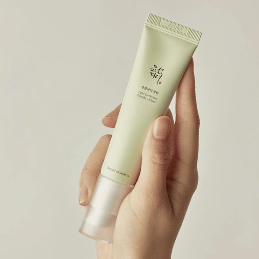A hand holds Beauty of Joseon Light On Serum with Centella and vitamin C, in a light green tube with minimalist design against a neutral background.