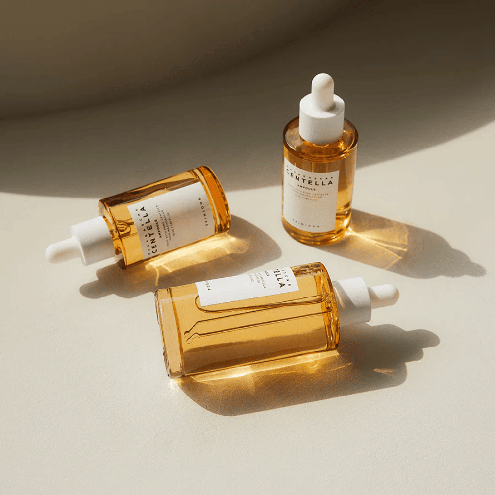 The image shows three bottles of "Madagascar Centella Asiatica 100 Ampoule". They are placed on a surface where sunlight creates a beautiful reflection through the golden contents of the bottles. The product appears to be part of a skincare series that uses centella asiatica, an ingredient known for its soothing properties. The design of the bottles is simple and modern, suggesting that it is a premium skincare series.