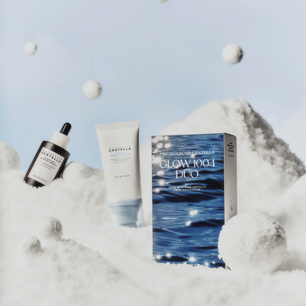The image shows the skincare set "Madagascar Centella Glow 1004 Duo" from SKIN1004, placed against a background depicting snow and sky.