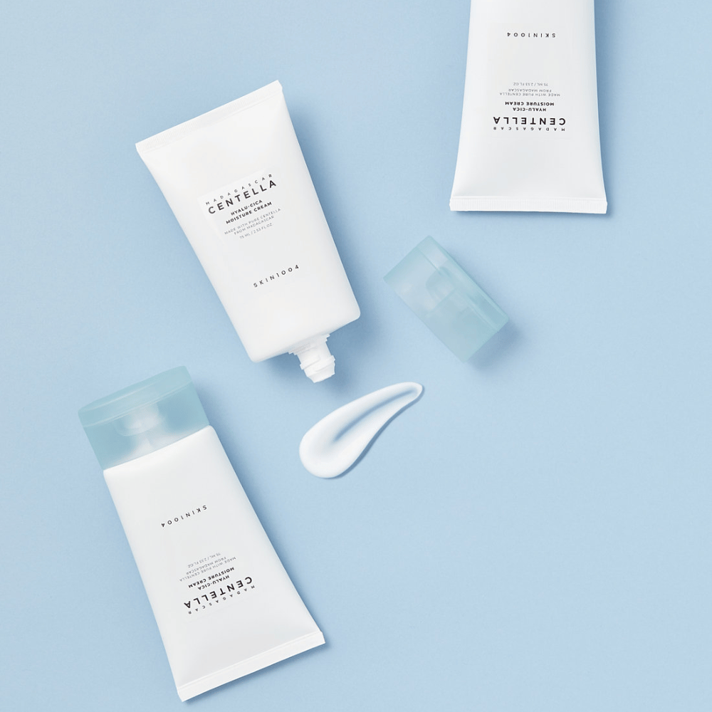 Two white tubes with SKIN1004 Centella skincare products, a dollop of product and a clear blue cap, on a light blue background.