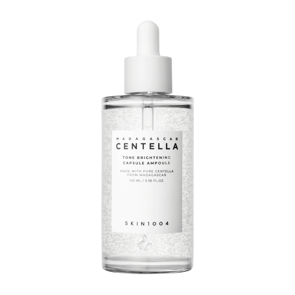 The image shows a bottle of "SKIN1004 Madagascar Centella Tone Brightening Capsule Ampoule". It is a transparent bottle with a white dropper applicator. The label states that the product contains pure centella ingredients from Madagascar. The contents of the bottle appear to have small capsules or particles, which may be part of its brightening skincare properties. The bottle contains 100 ml / 3.38 fl.oz. of the product.