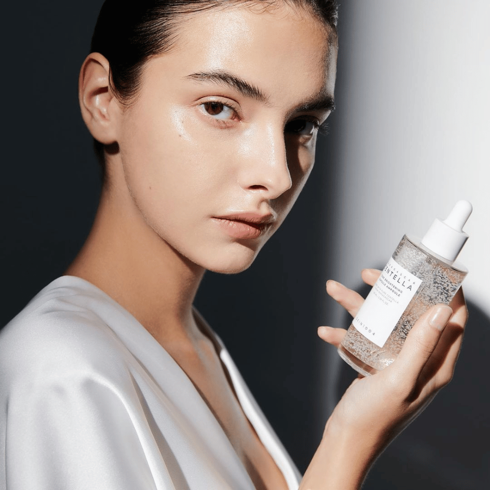 In the image, a person is seen holding a bottle with a dropper applicator. The bottle is labeled "SKIN1004 Madagascar Centella Tone Brightening Capsule Ampoule". The person has a shimmering skin tone that reflects the light, suggesting a possible effect of the product.