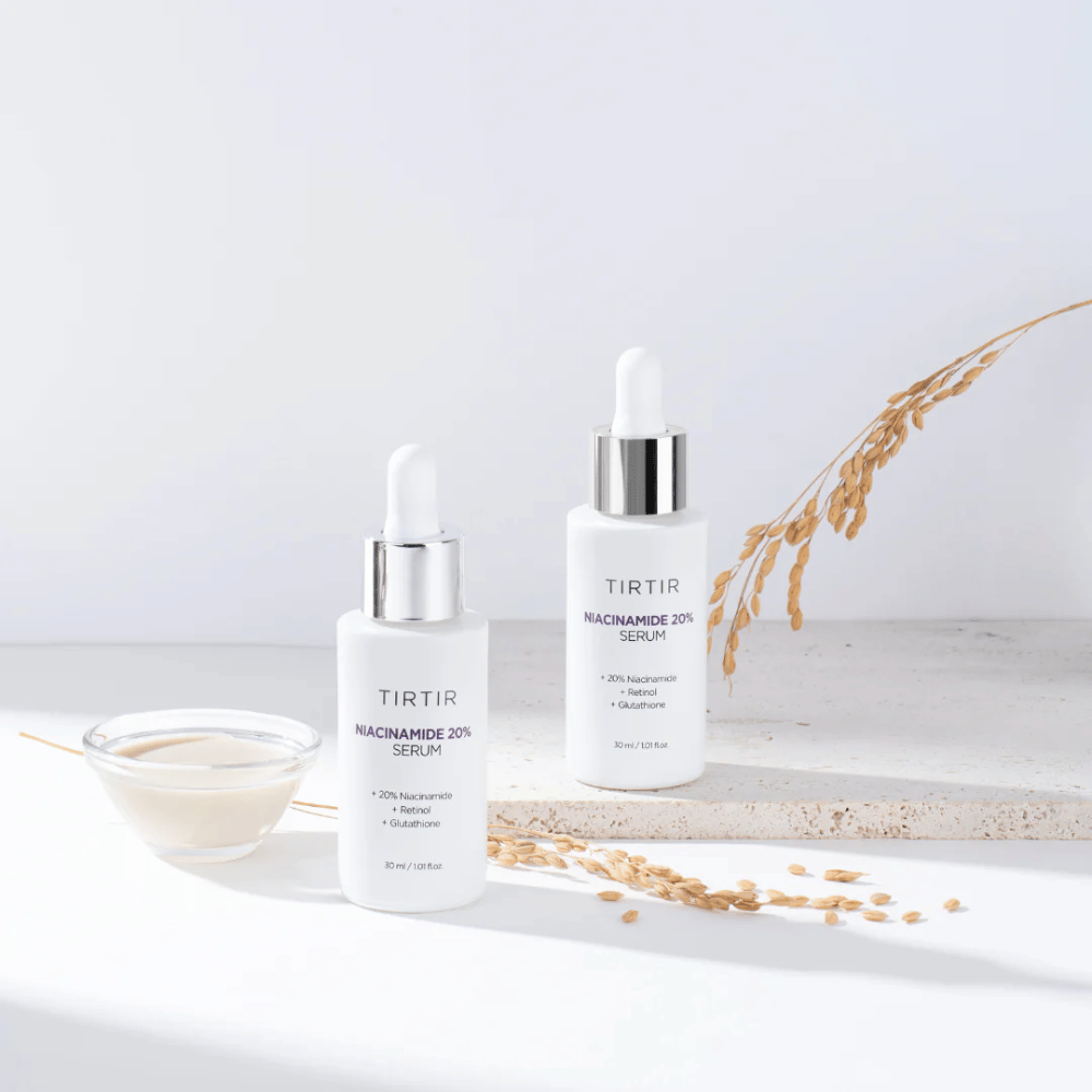 Two bottles of Tirtirs "Niacinamide 20% Serum" are displayed on a light surface along with a small bowl of a creamy substance and some strands of dry plants. The serum contains 20% niacinamide, retinol, and glutathione, which helps to improve the skin's appearance and texture.