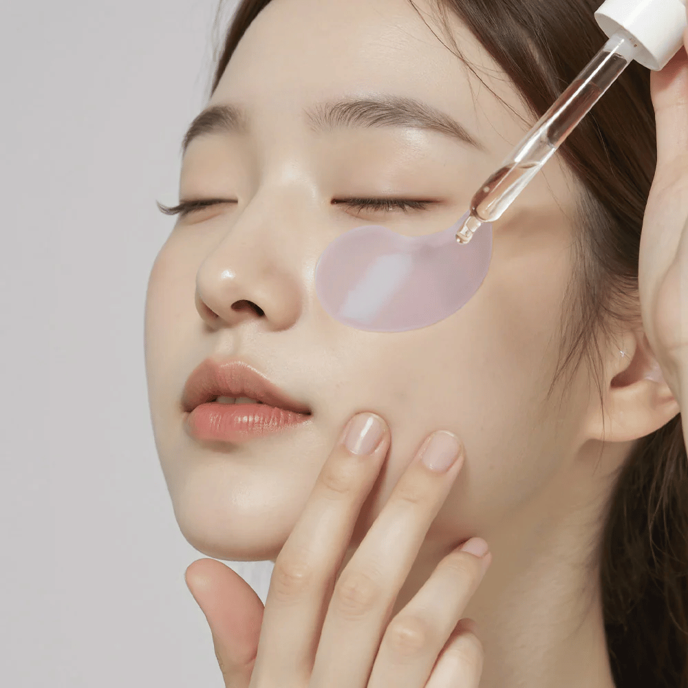 A woman with a purple hydrogel eye patch applies serum under the eye with a pipette. The product moisturizes and cares for the sensitive skin around the eyes.
