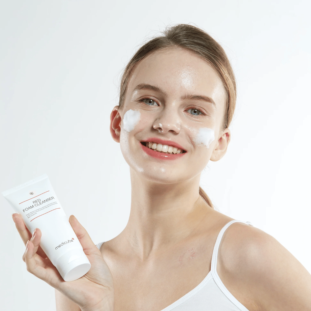 Young woman with cleansing foam on her face holding up a tube of Medicube Red Foam Cleanser and smiling at the camera. She is wearing a white tank top and the background is light and simple.